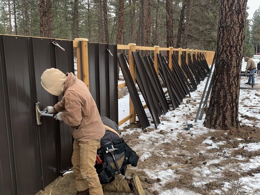Crew installing black metal panels on a fence in a snowy forest